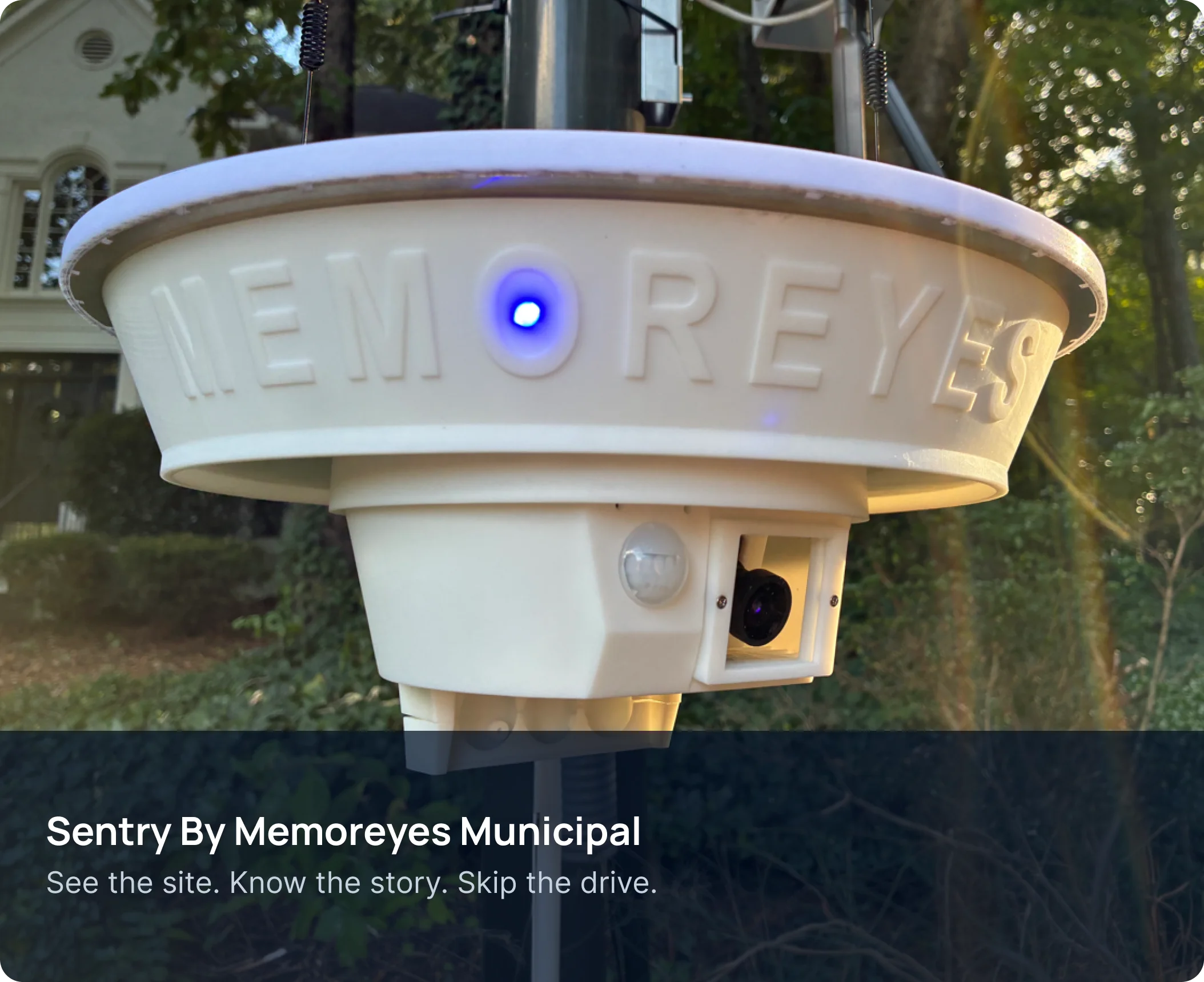 A white, round, outdoor security camera with a blue light on its front, mounted on a pole in front of a house with trees in